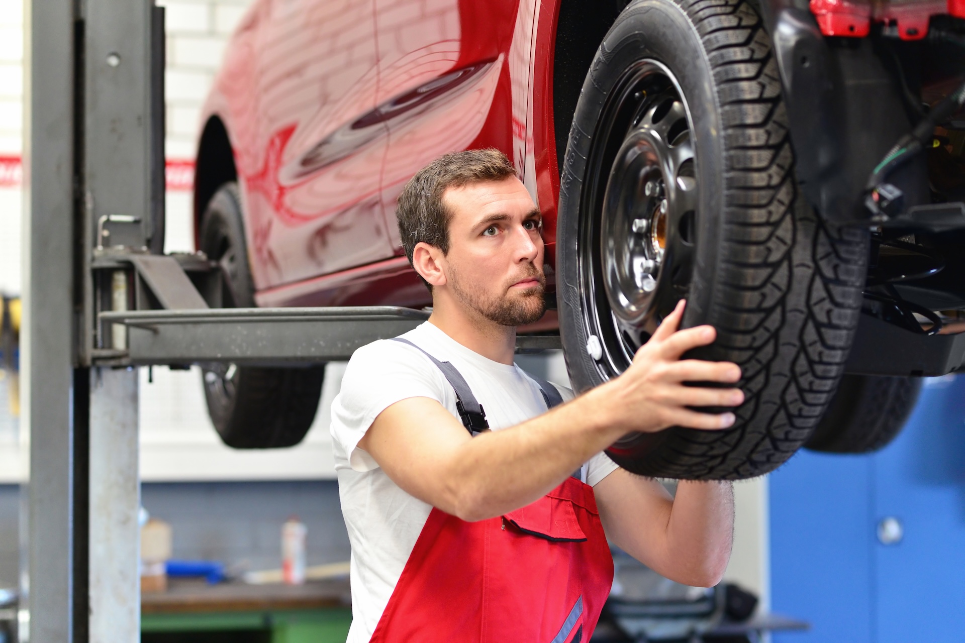 Auto mechanic in red overalls installing a tire on a red car lifted on a hydraulic platform in a professional garage.