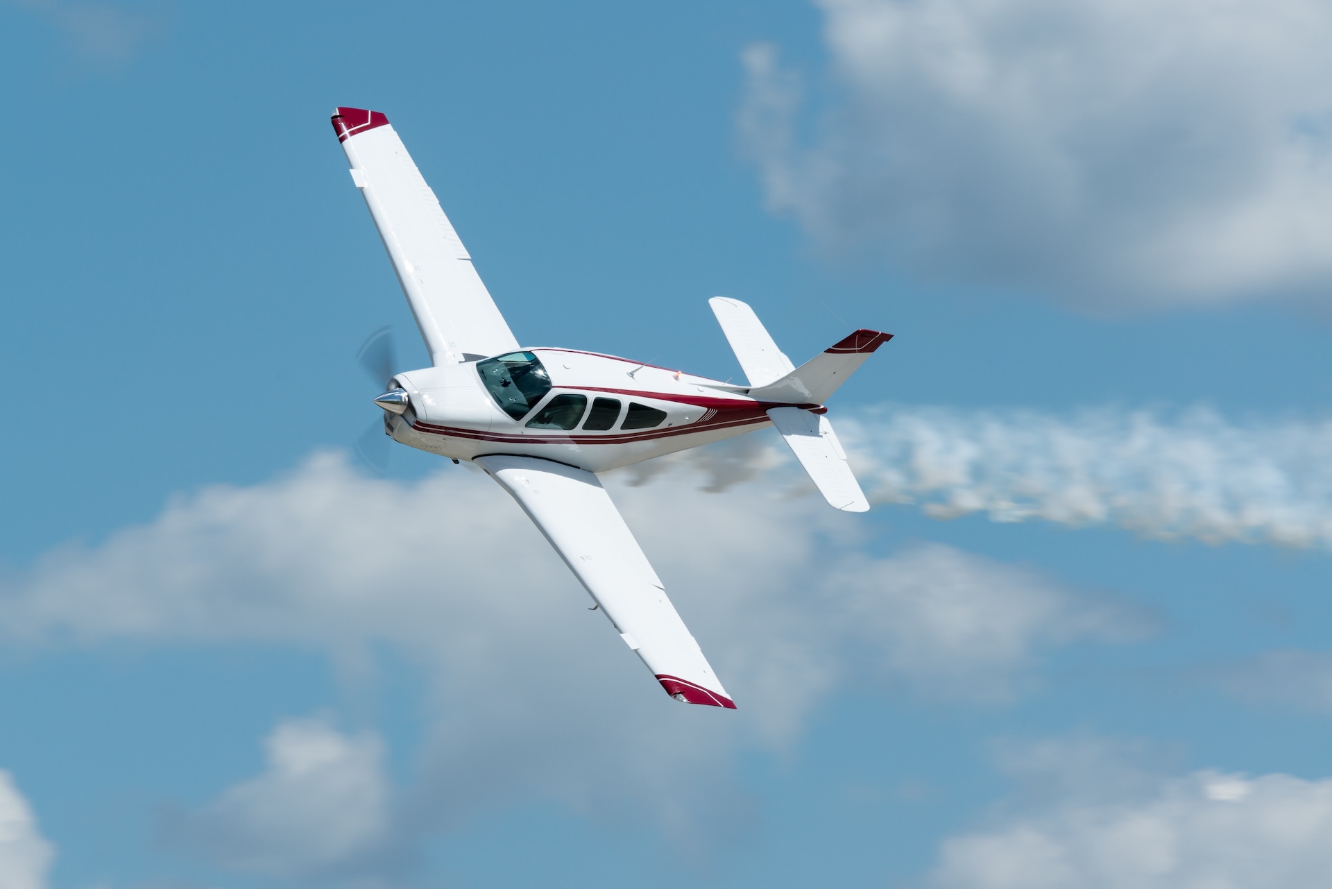 White and red single-engine airplane performing an aerobatic maneuver with a smoke trail against a blue sky with clouds.