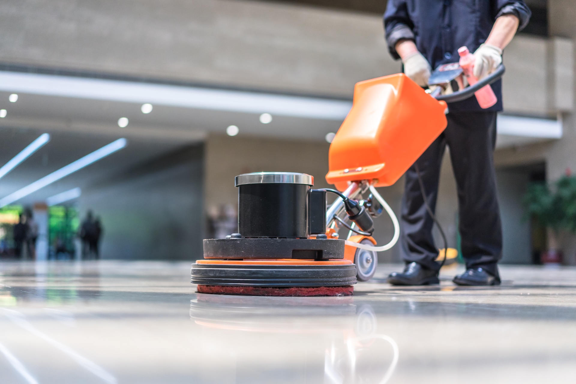 Close-up of an orange industrial floor scrubber being operated by a person in black attire, cleaning a shiny hallway floor in a modern building.
