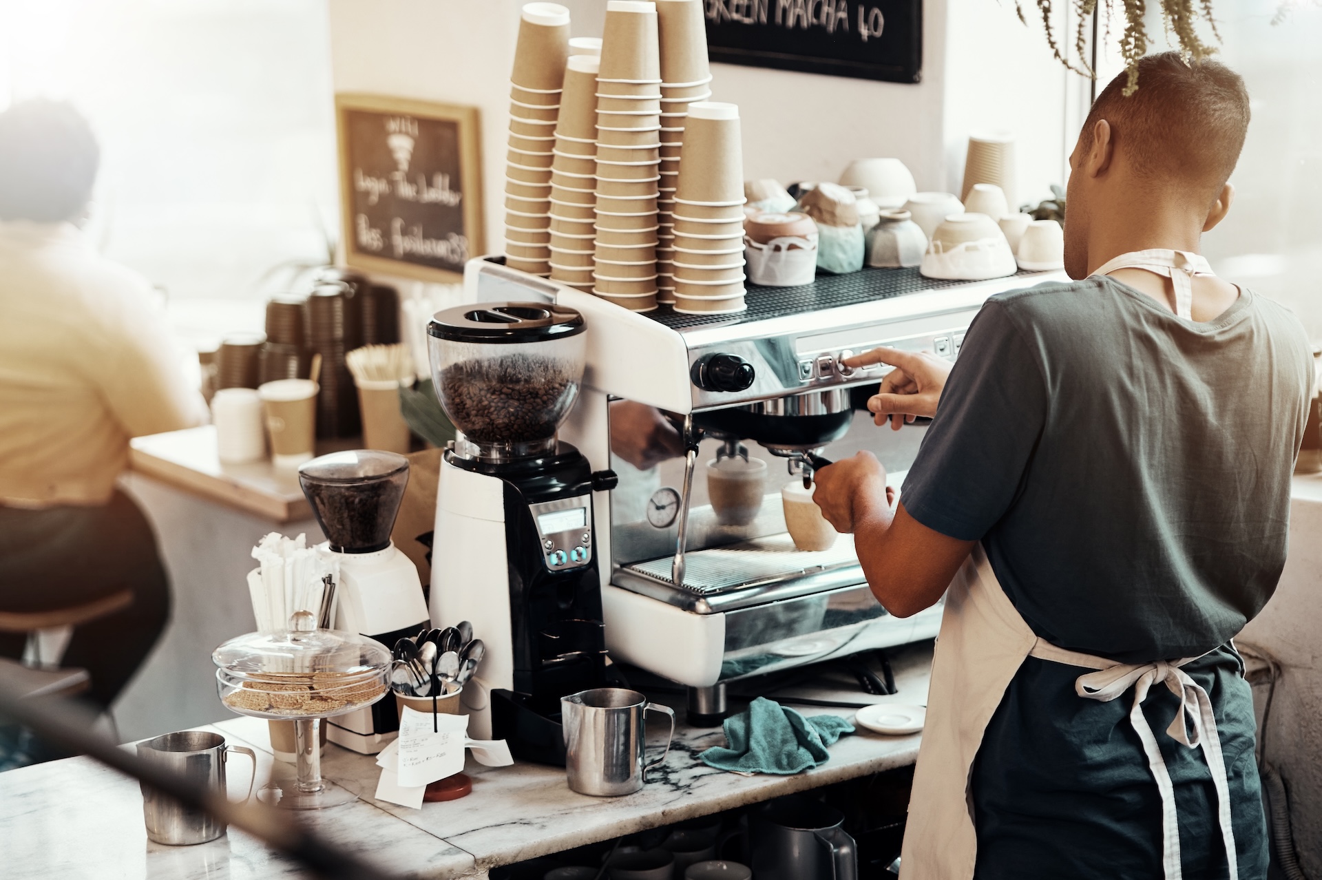 Barista using an espresso machine with paper cups and jars in the foreground.