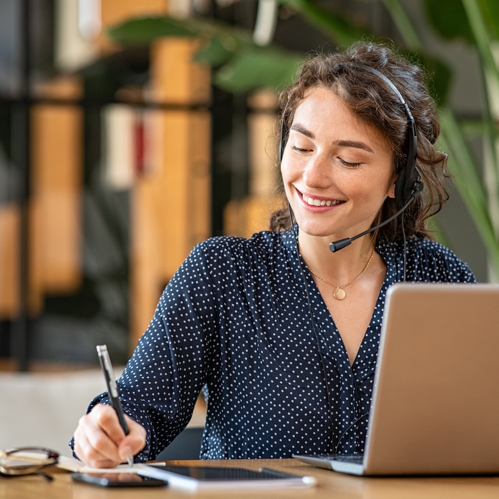 a smiling woman working at her desk with a headset and laptop, taking notes with a pen