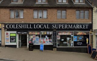 Front view of Coleshill Local Supermarket with various signs in the windows and a visible 'Welcome' sign above the entrance.