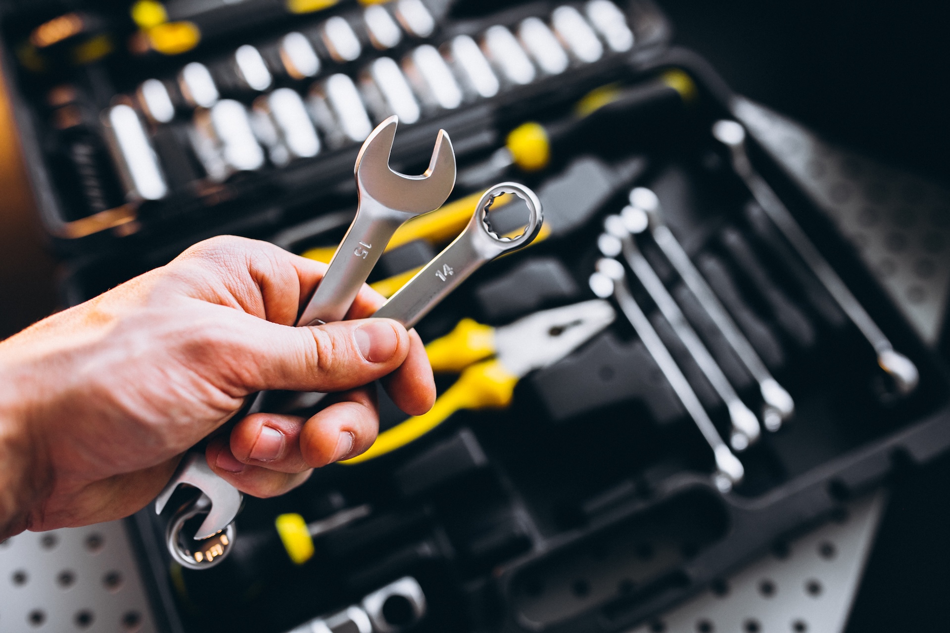 Hand holding two silver wrenches above an open toolset with yellow accents.