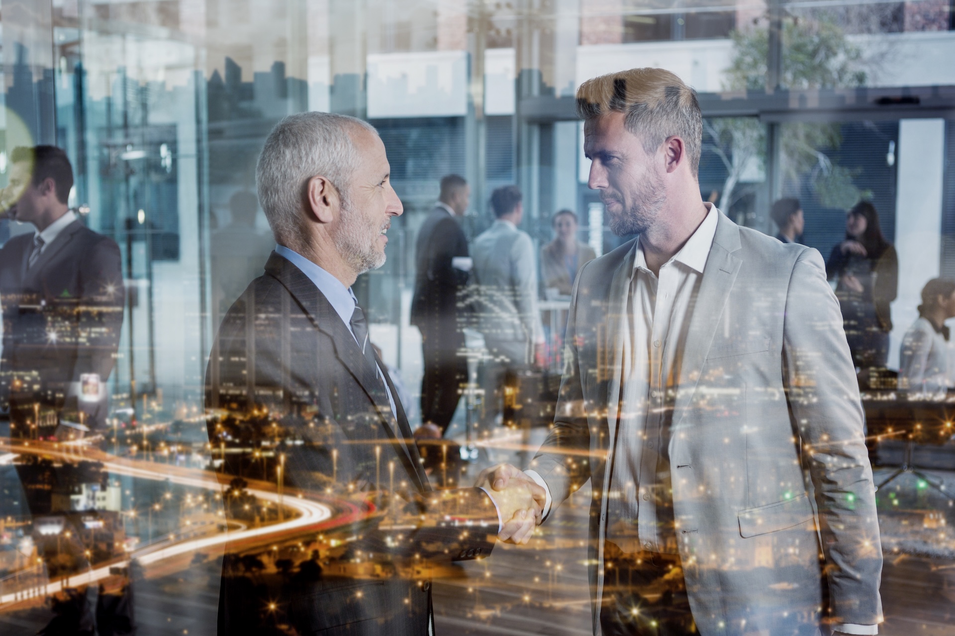Two men shaking hands in a modern office setting with a night cityscape overlay, representing a successful business deal or acquisition.