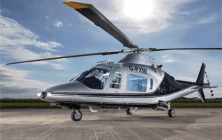 High-resolution image of a silver and black helicopter marked G-PXMI, sitting on an airstrip with the sun shining overhead.