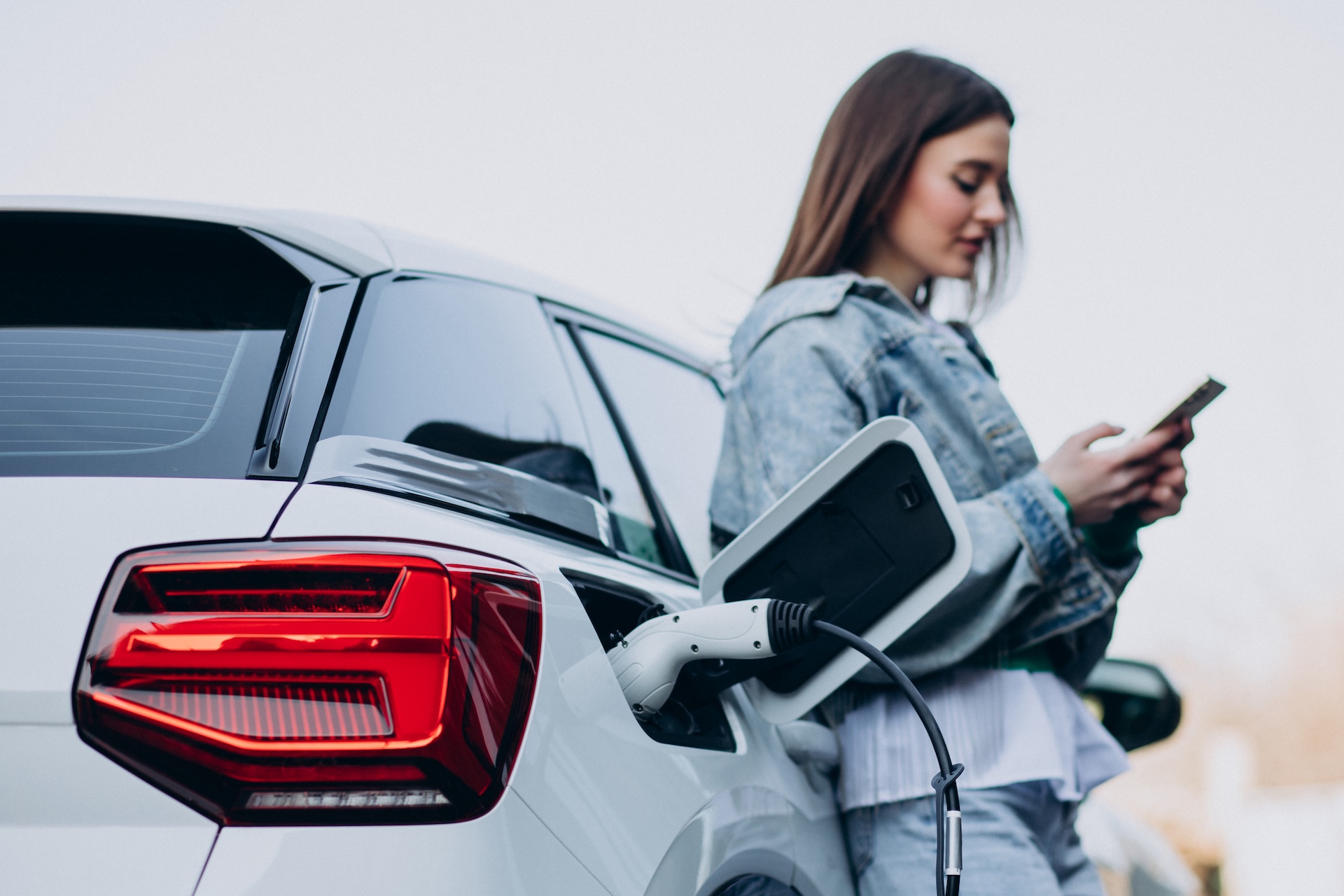 Woman charging her electric car with charging pistol A image of a woman resting up against the drivers side of her car, using her phone wihlie charging her electric car