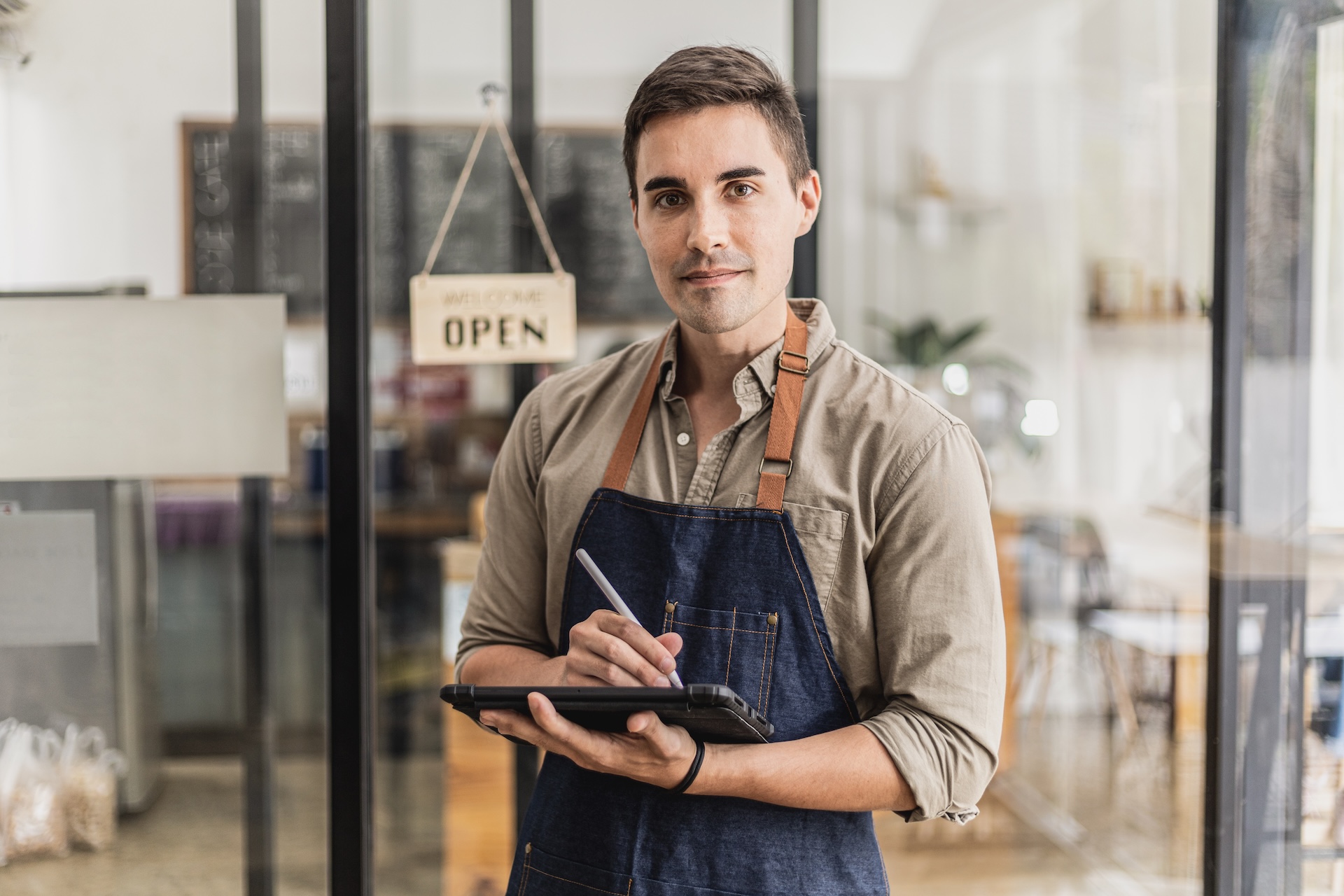 Young male store clerk in an apron holding a tablet inside a cafe, taking notes or serving customers.