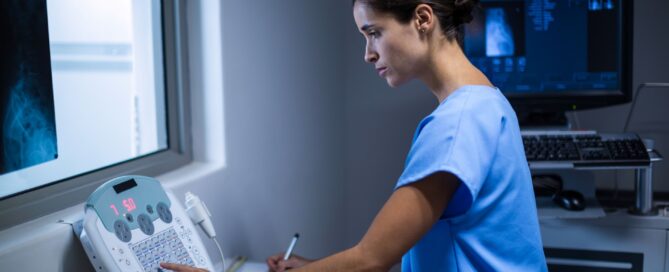 Full version of a female technician using an X-ray machine and taking notes in a darkened diagnostic room.
