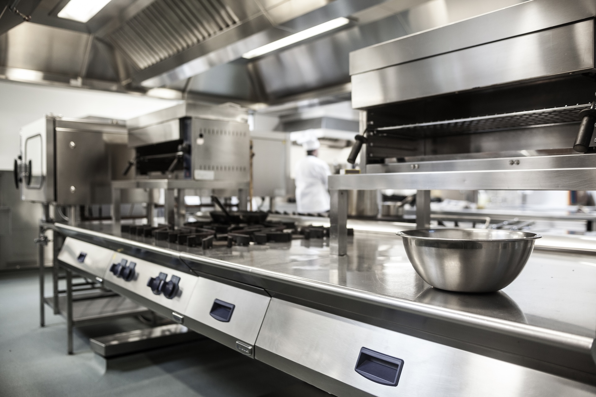 A image taken in a professional kitchen showing silver worktops, cookers, grills and a silver bowl