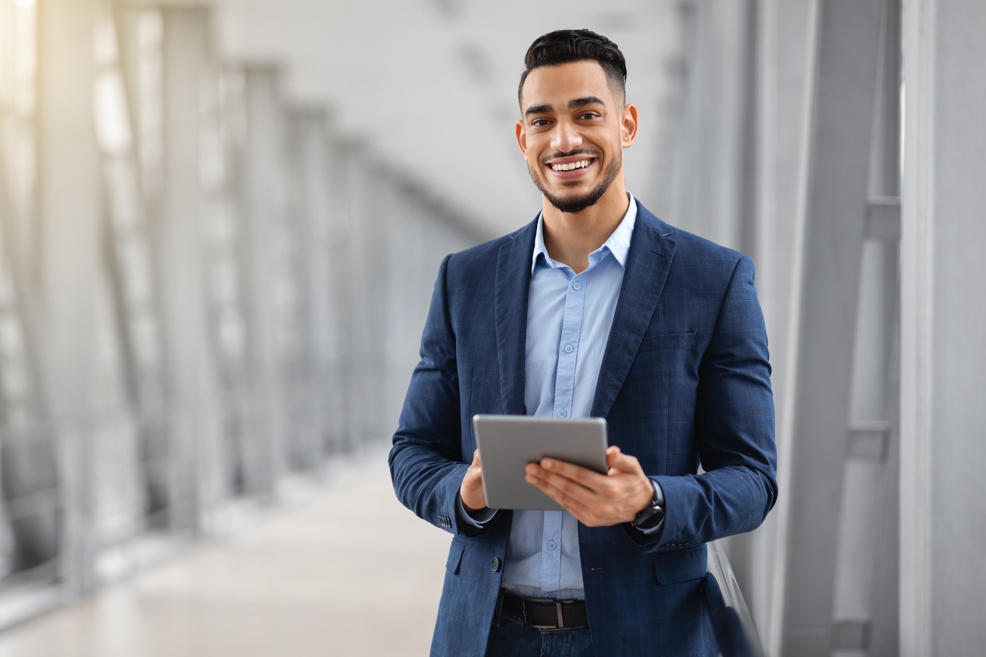 Smiling,Young,Middle,Eastern,Man,With,Digital,Tablet,In,Hands A businessman in a dark suit smiles while using a mobile phone, standing in an office environment.