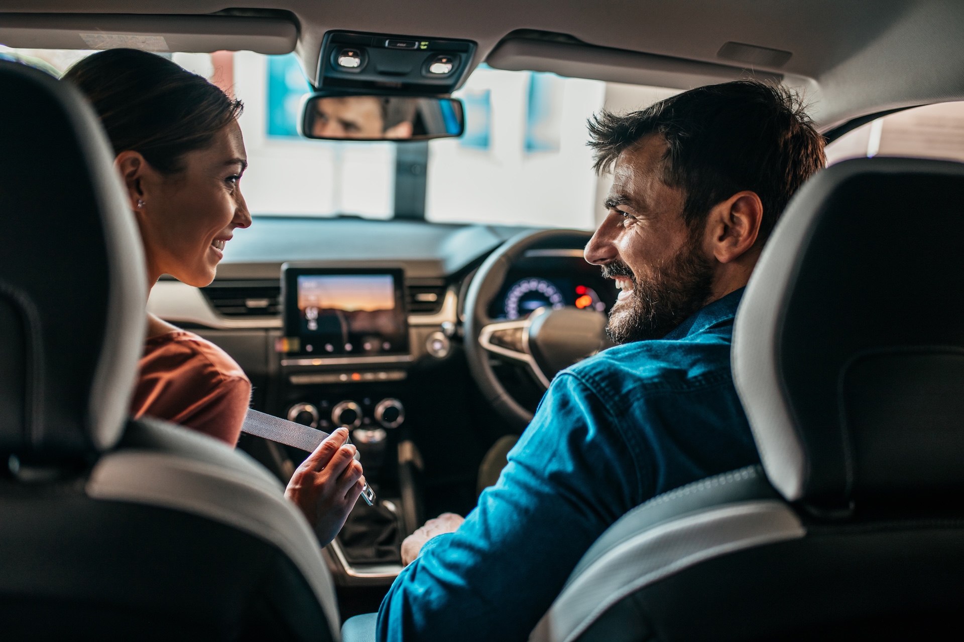 A image taken from the back seat of a car showing a man and woman smiling at each other in the front seats